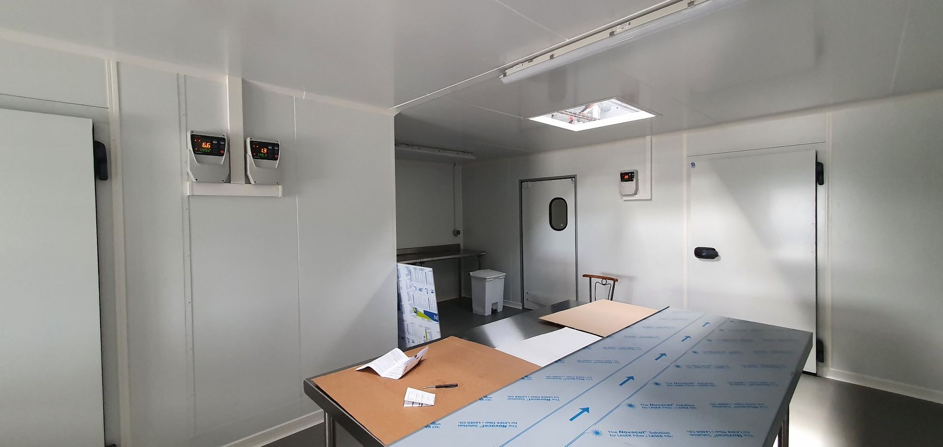 Vue intérieure d'une cuisine commerciale blanche avec tables, portes et éclairage suspendu en acier inoxydable.