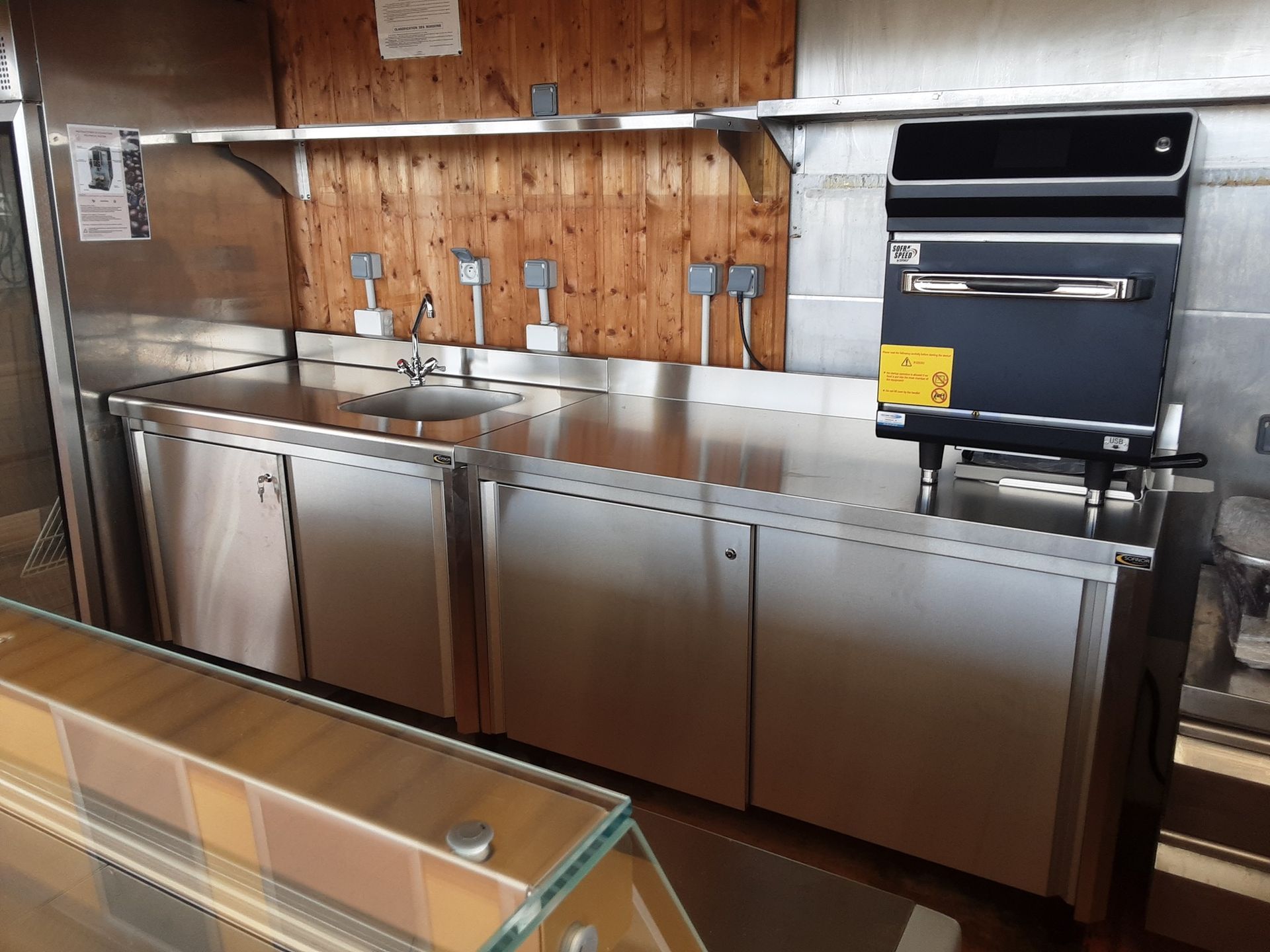 Comptoir de cuisine commerciale en acier inoxydable avec évier, armoires et four, adossé à un mur lambrissé.