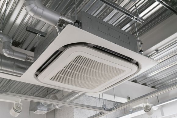 Unité de climatisation/chauffage fixée au plafond, avec panneau blanc et conduits métalliques. Observée dans un bâtiment commercial ou industriel.