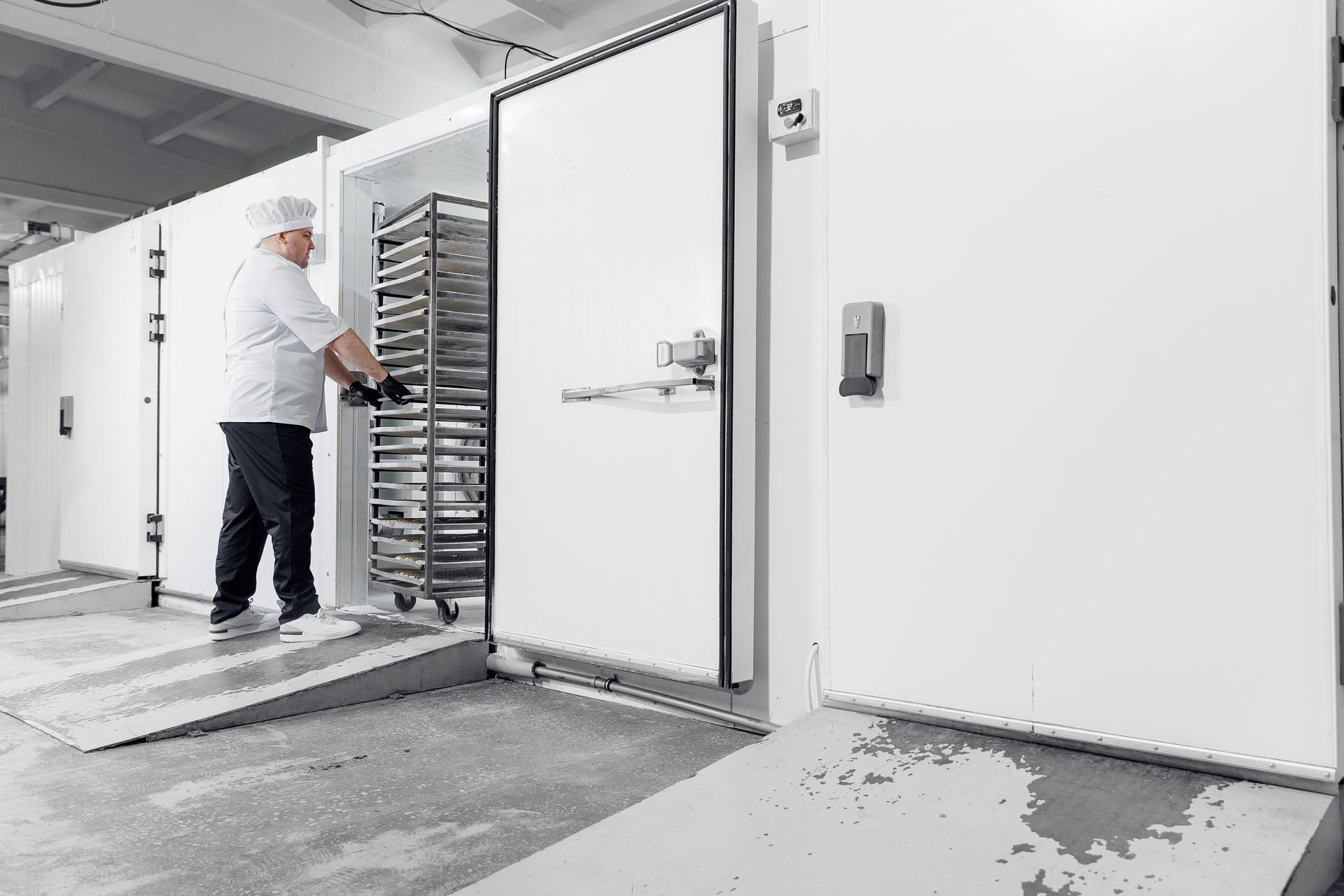 Une personne en uniforme blanc pousse un chariot de plateaux dans une grande chambre froide blanche.