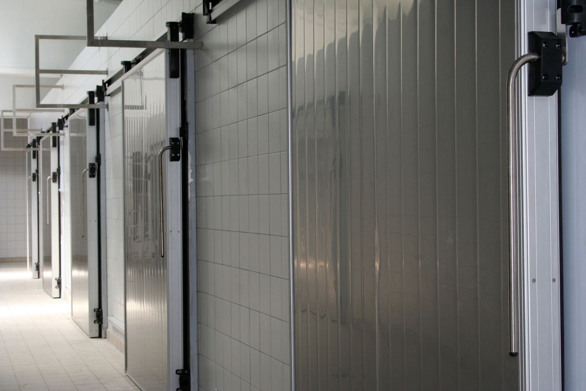 Des portes coulissantes métalliques alignées donnent accès à des chambres froides, aux murs carrelés et lambrissés de blanc.