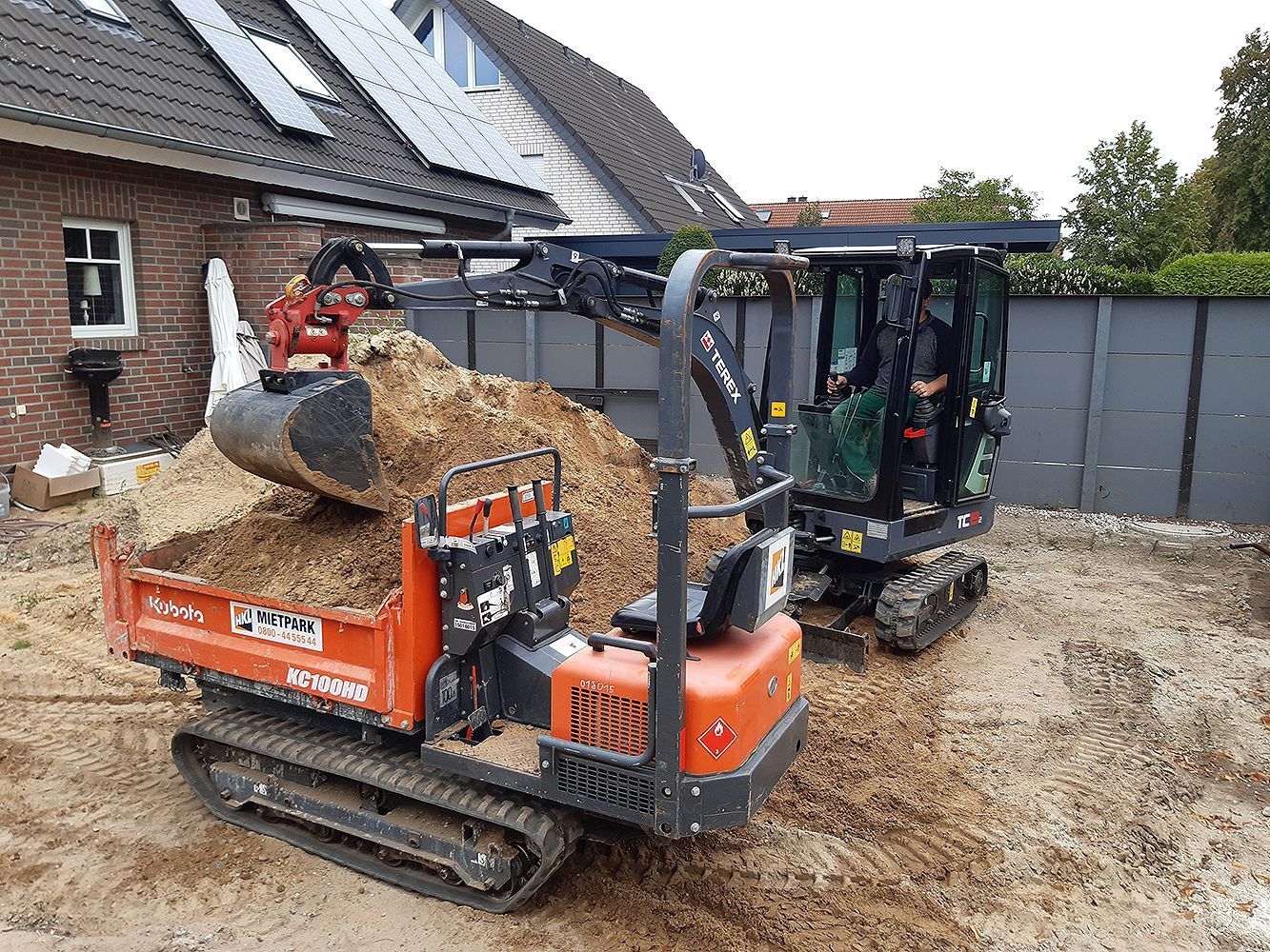 Ein kleiner Bagger lädt Erde in einen Müllcontainer.