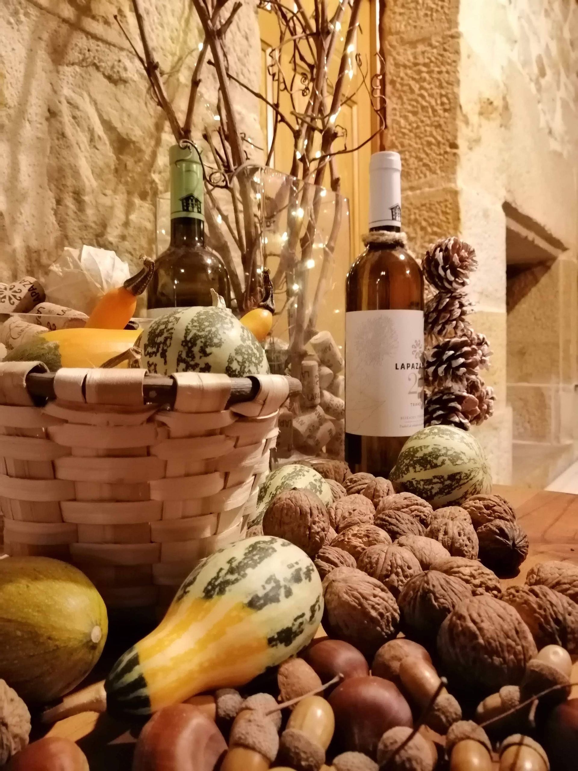 Bodegón de calabazas, nueces y botellas de vino; cesta y ramitas sobre superficie de madera.