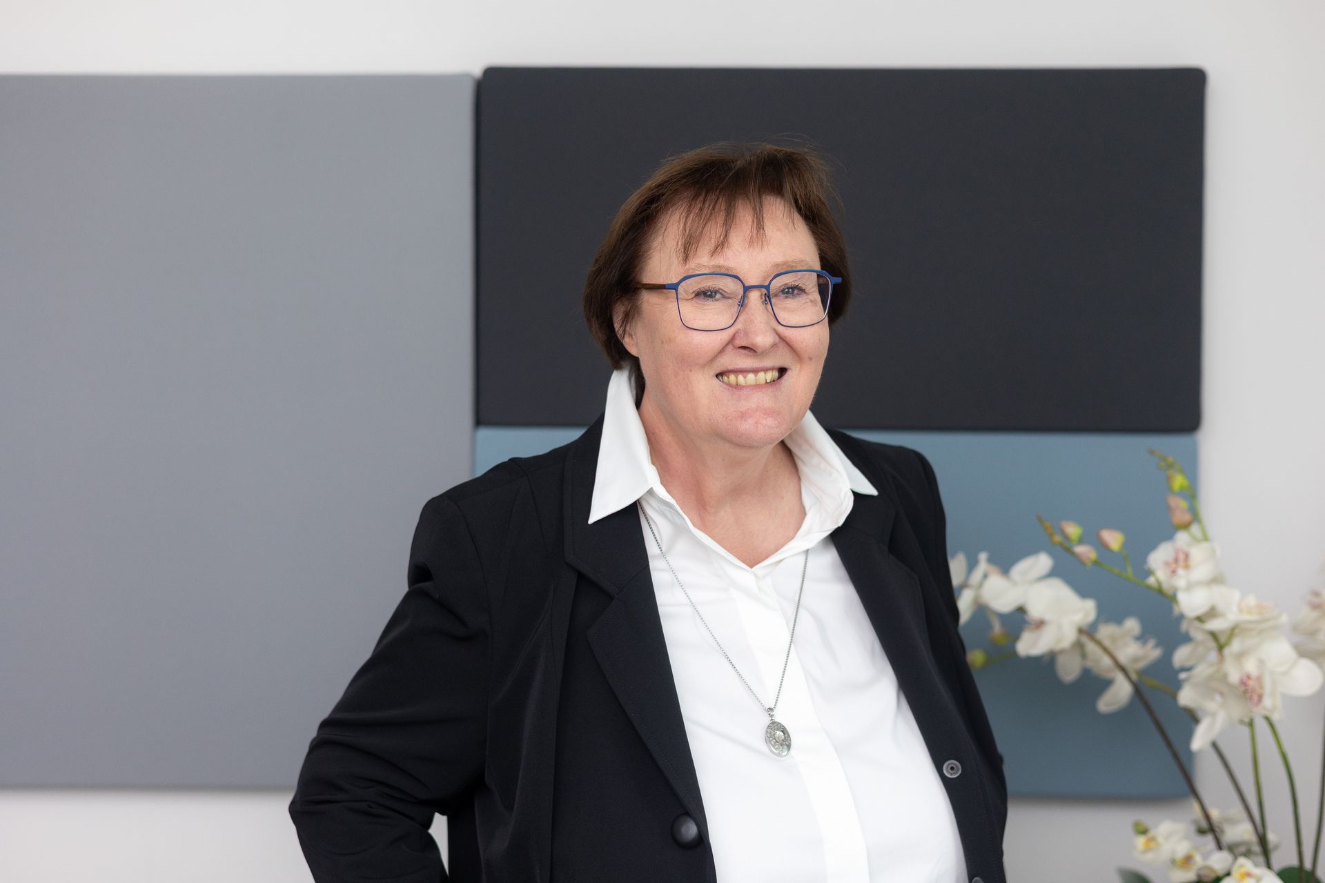 Person mit schwarzem Blazer, weißem Hemd und silberner Halskette vor grauen und schwarzen Wandpaneelen.