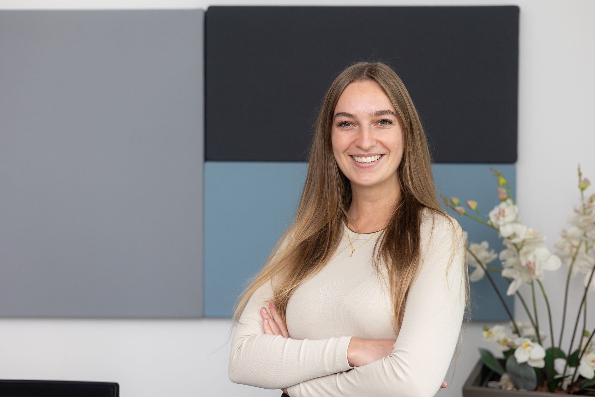 Frau mit langen braunen Haaren, weißem Langarmshirt und verschränkten Armen vor einer Wand.