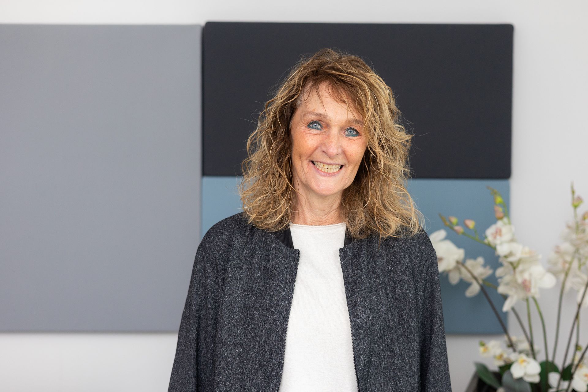 Frau mit schulterlangem, lockigem, blondem Haar trägt weißen Pullover und dunkelgraue Strickjacke.