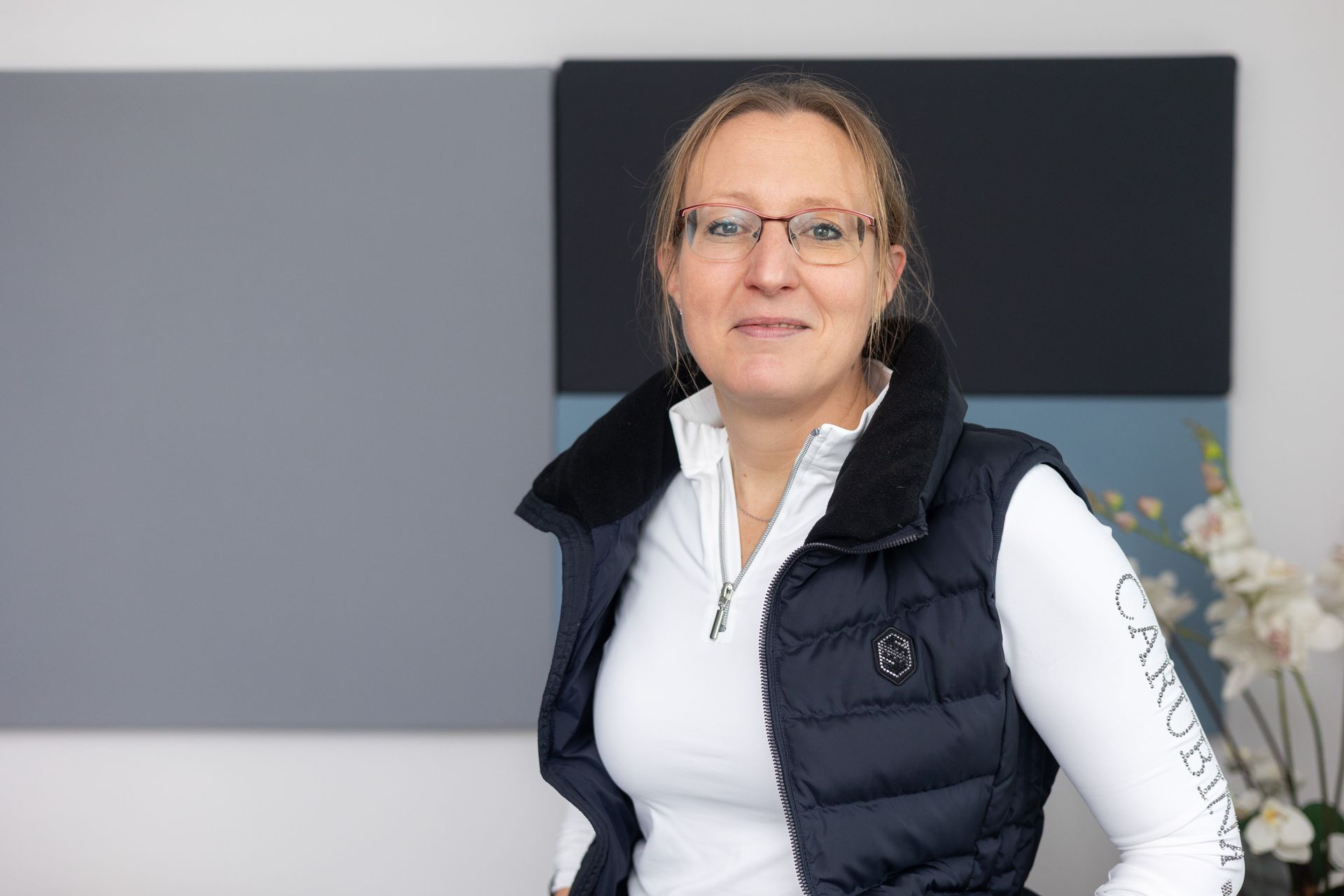 Person mit blondem Haar trägt weiße Langarmshirt mit Reißverschluss und schwarzer Steppweste vor grau-blauen Wandpaneelen.