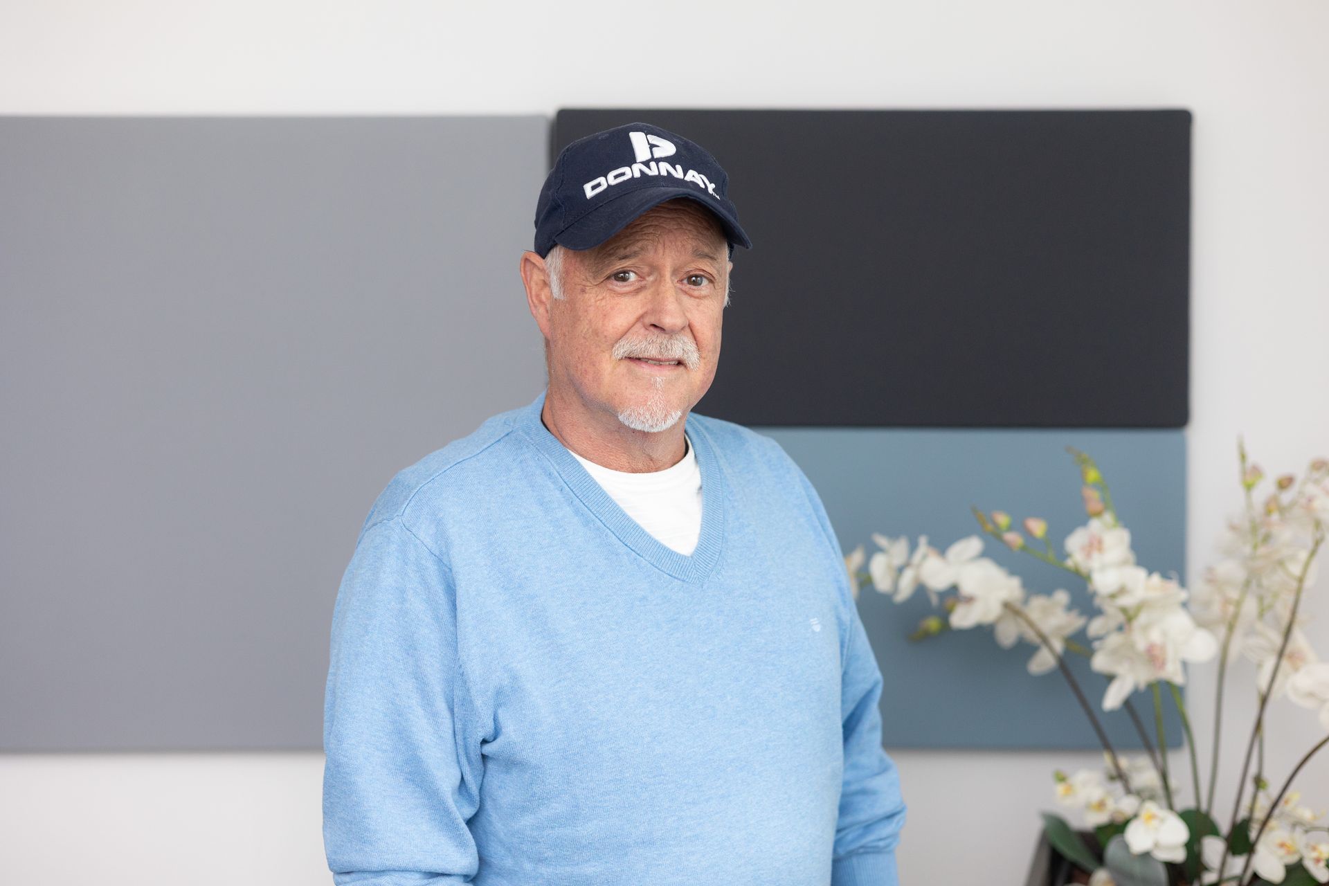 Mann mit blauem Pullover und dunkelblauer Kappe mit weißem Schriftzug 'DONNAY' vor grauen Wandpaneelen und Orchideen.