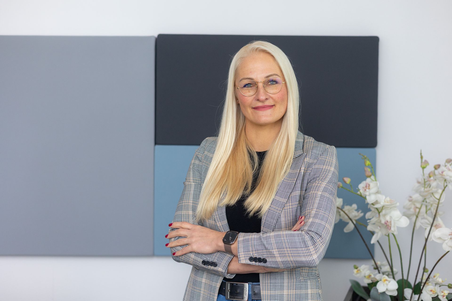 Frau mit langen blonden Haaren, kariertem Blazer und schwarzem Oberteil steht mit verschränkten Armen.