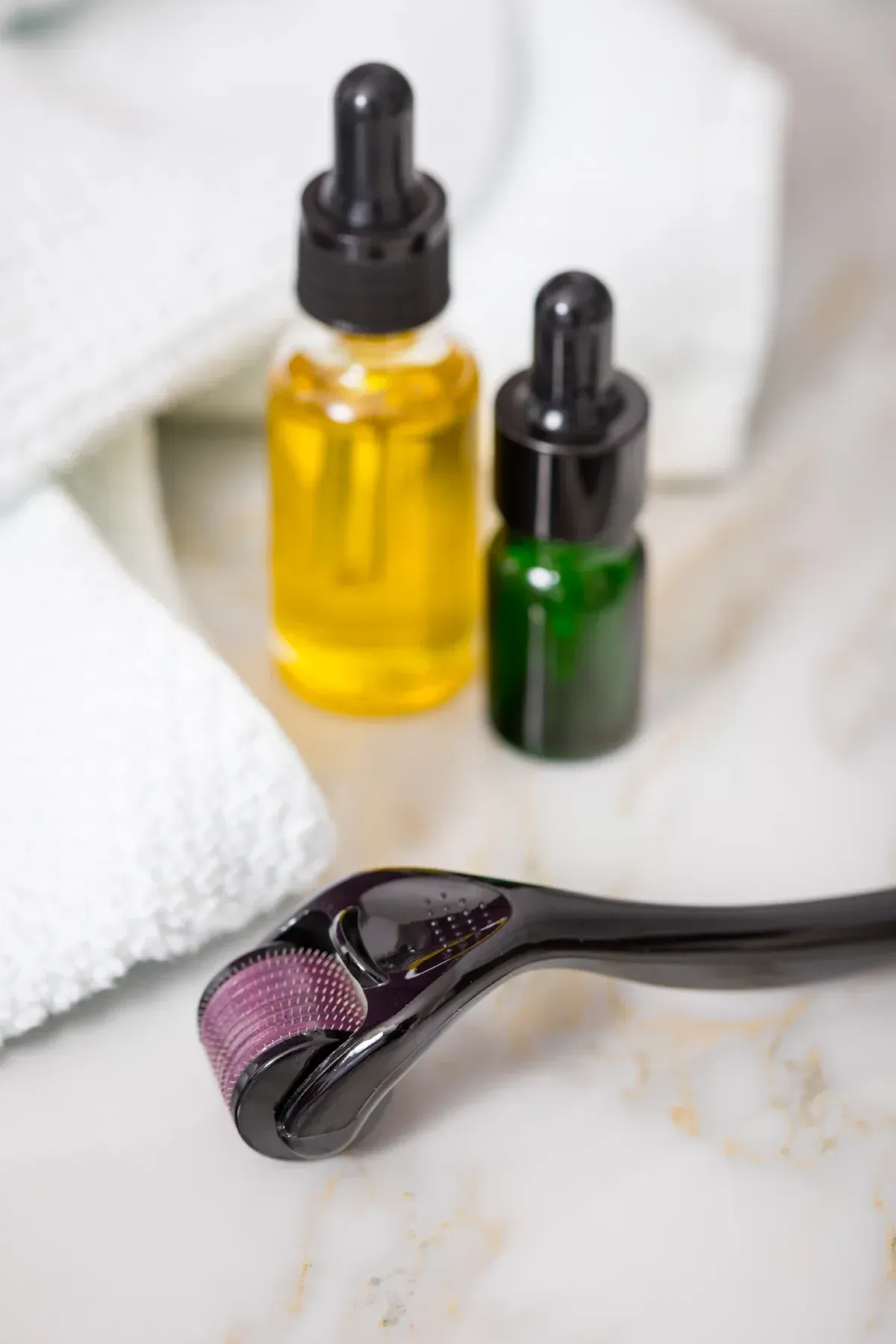 Rodillo de microagujas con frascos goteros de aceite sobre una superficie de mármol blanco, junto a toallas.
