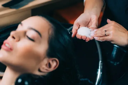 Mujer lavándose el pelo en la peluquería. Se aplica champú con las manos, con los ojos cerrados, en un lavabo negro.
