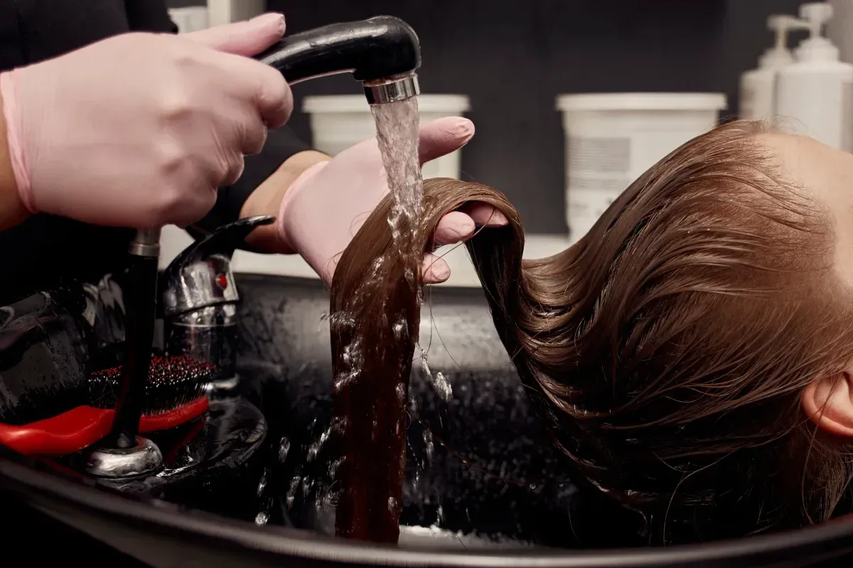 Peluquera lavando el cabello de un cliente en el lavabo del salón; agua cayendo sobre el cabello castaño
