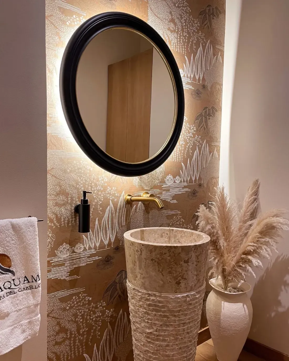 Tocador con espejo redondo, lavabo de piedra, grifo dorado, papel tapiz con estampado beige y pasto de las pampas.