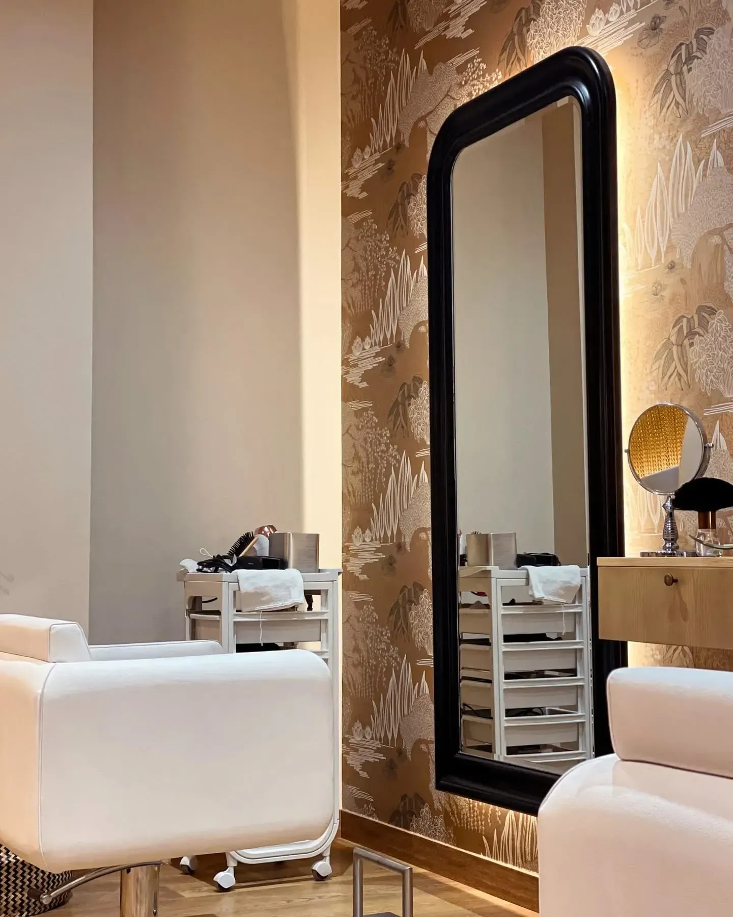 Interior de peluquería con un gran espejo con marco negro, sillas blancas y un carrito con suministros.