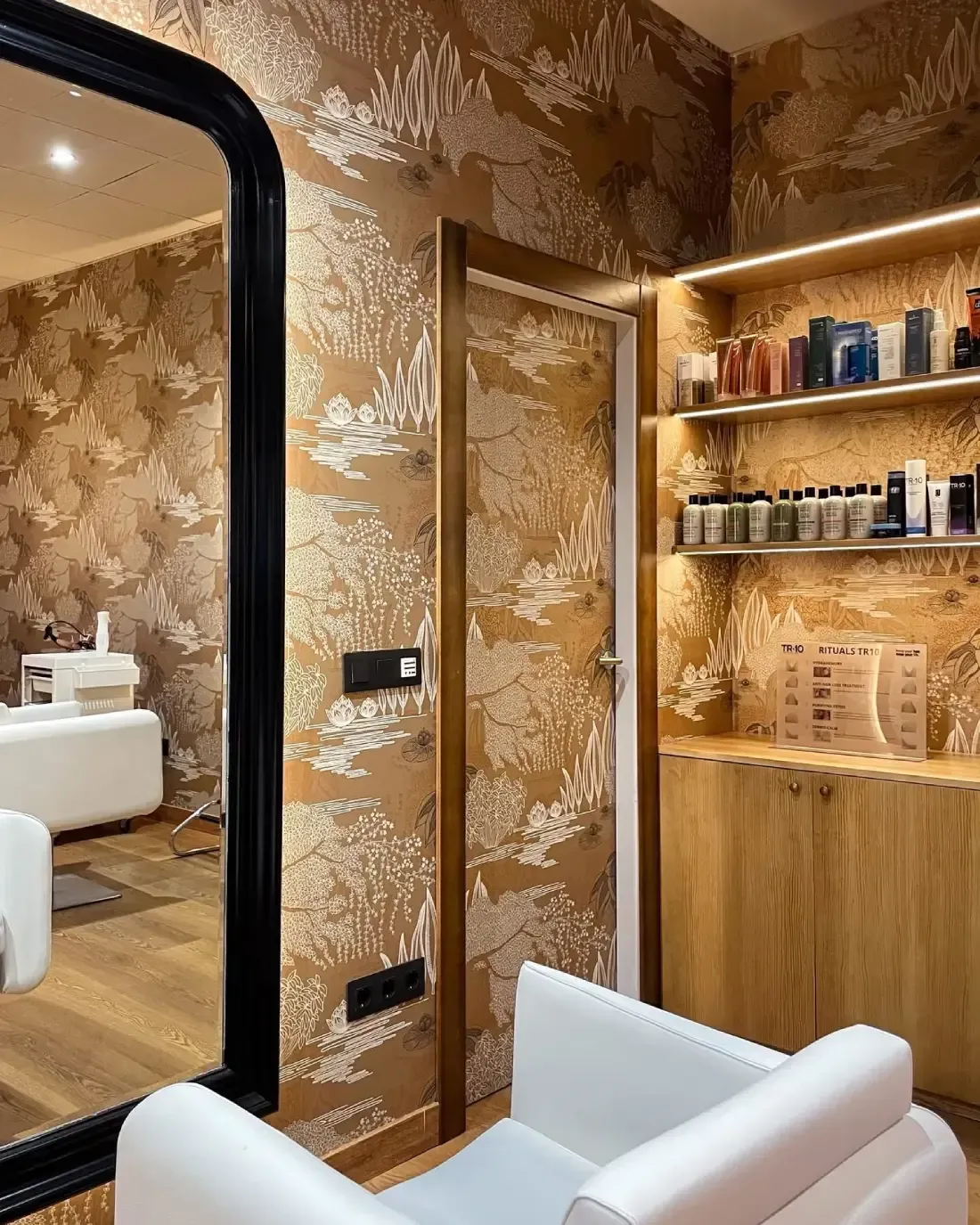 Interior de salón con papel tapiz estampado, espejo grande, silla blanca y estantes para productos.