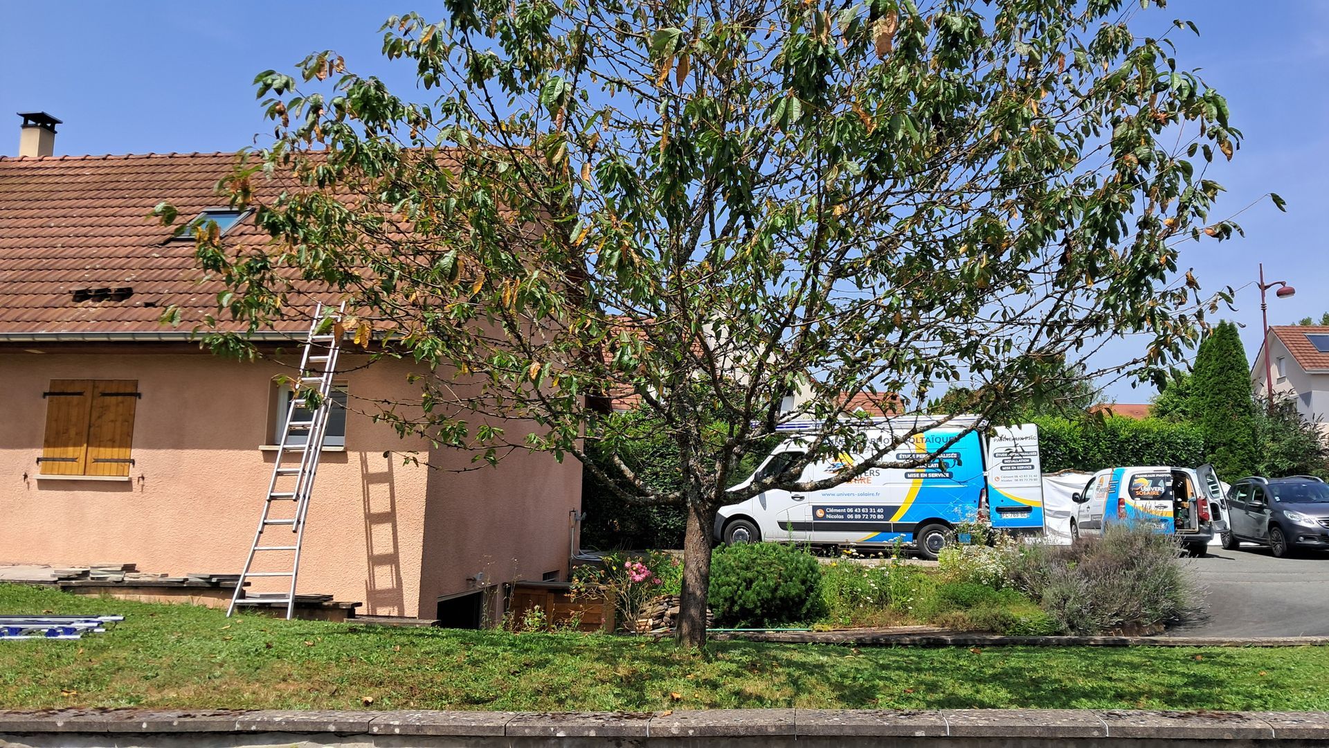 Photo d'une maison rose sur la gauche avec une échelle allant sur le toit. Il y a un arbre au milieu de la photo et les camions de l'entreprise sur la droite