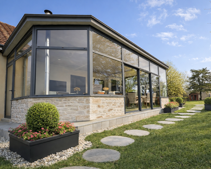 Espace repas extérieur couvert avec toit en verre et portes coulissantes en verre, table et chaises en bois.