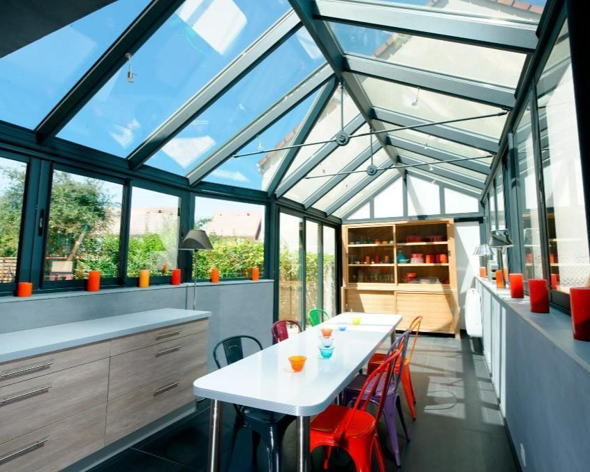 Véranda avec toit et fenêtres en verre, table et chaises, placards de cuisine. Espace lumineux et ouvert.