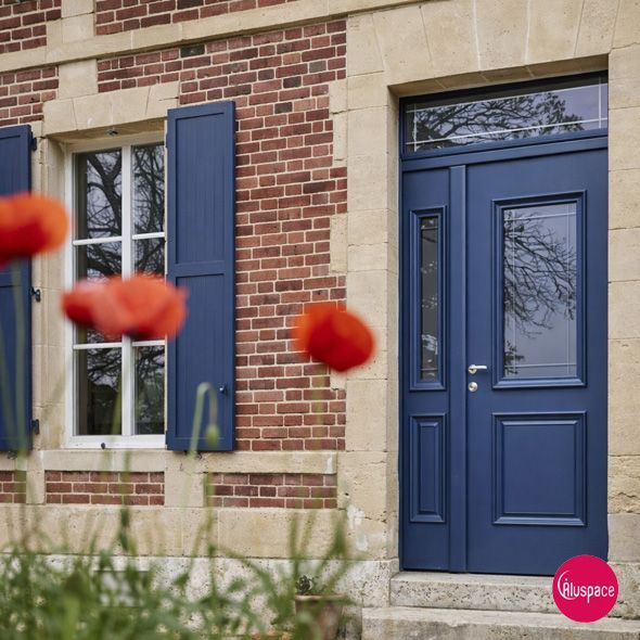 Porte d'entrée bleue avec imposte et fenêtre latérale sur un bâtiment en briques avec volet bleu.