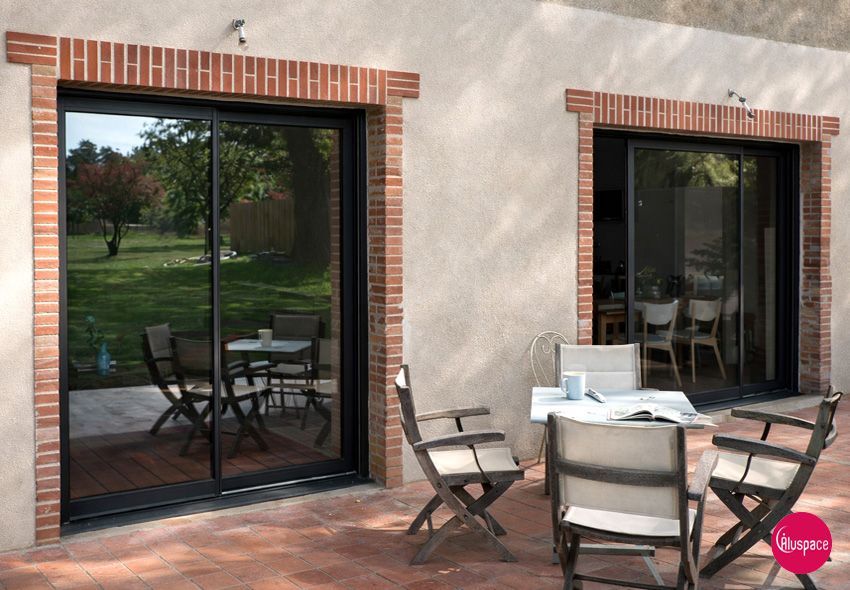 De larges portes coulissantes en verre s'ouvrent sur une terrasse aménagée avec une table, des chaises et une piscine. Encadrements noirs, façade lumineuse.