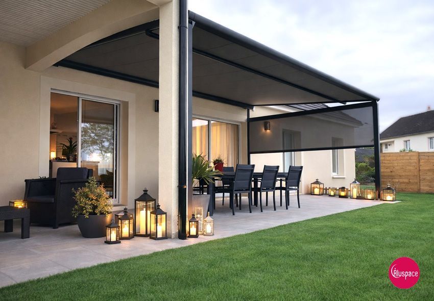 Auvent noir au-dessus d'une terrasse avec table et chaises noires, maison beige et petite clôture en piquets.