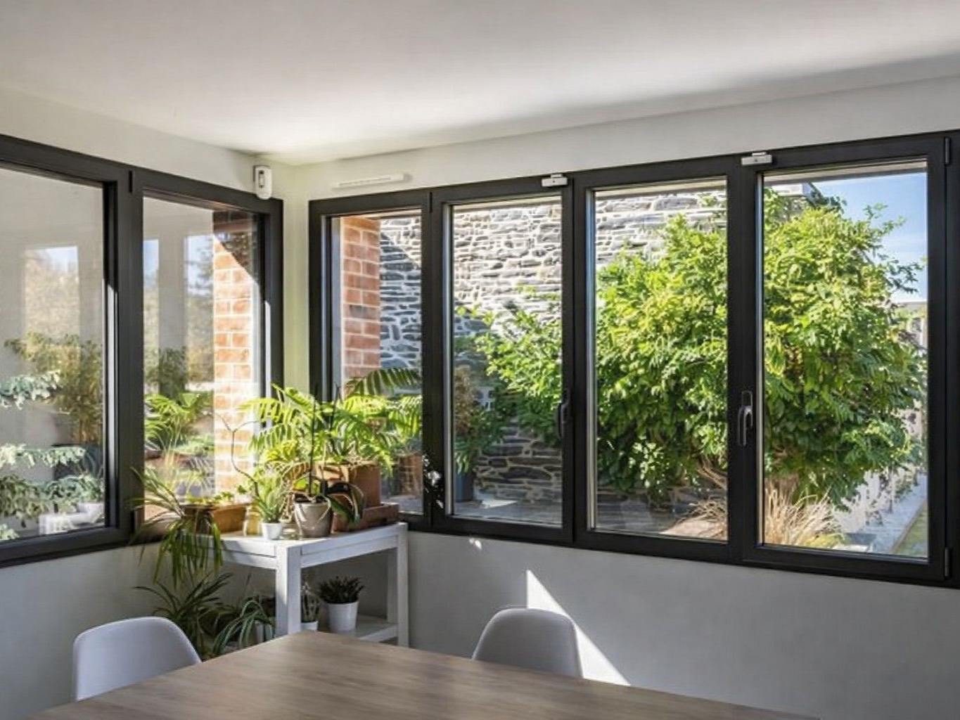 Véranda aux fenêtres à cadre noir, baignée de lumière. Vue sur la verdure extérieure. Table et chaises au premier plan.