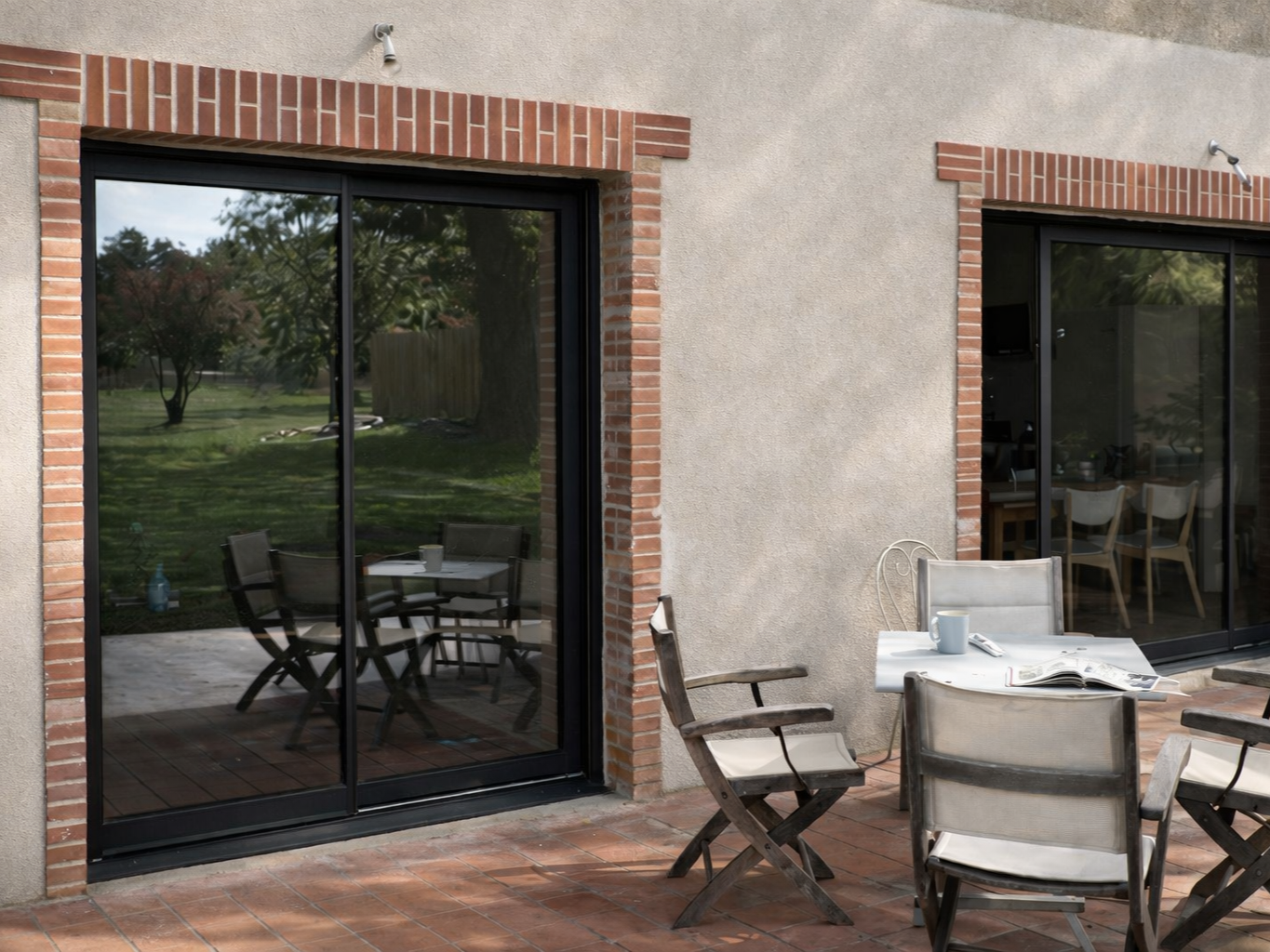 De larges portes coulissantes en verre s'ouvrent sur une terrasse aménagée avec une table, des chaises et une piscine. Encadrements noirs, façade lumineuse.