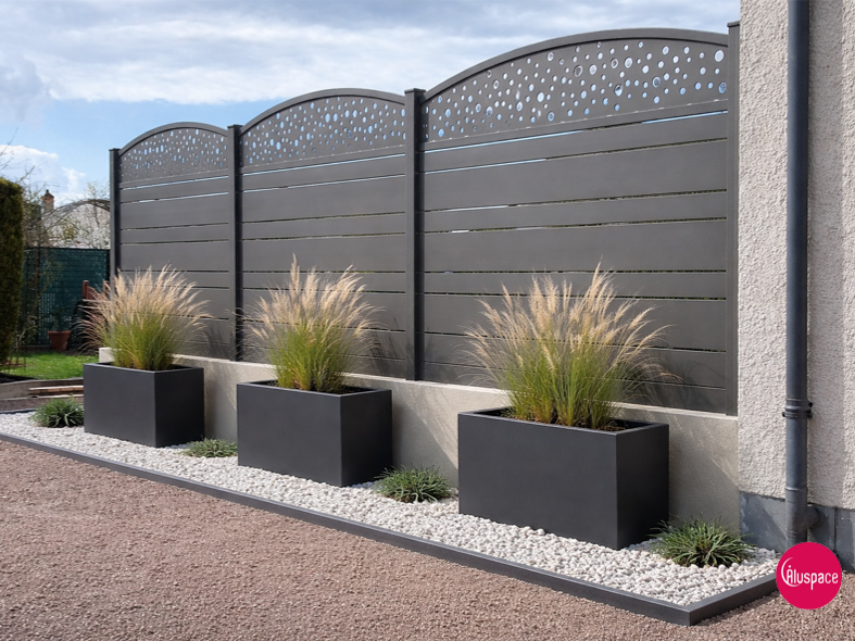 Clôture à lattes horizontales grises avec partie supérieure décorative, adossée à une terrasse en briques et à un arbre en fleurs.