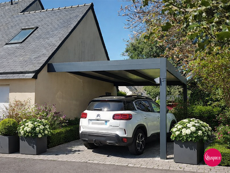 Une voiture argentée est garée sous un abri de voiture gris. Pelouse verte, maison en arrière-plan.