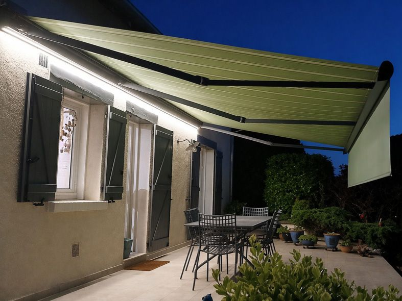 Un store vert rétractable protège une terrasse du soleil au crépuscule, tandis que des lumières illuminent la maison et les meubles en contrebas.