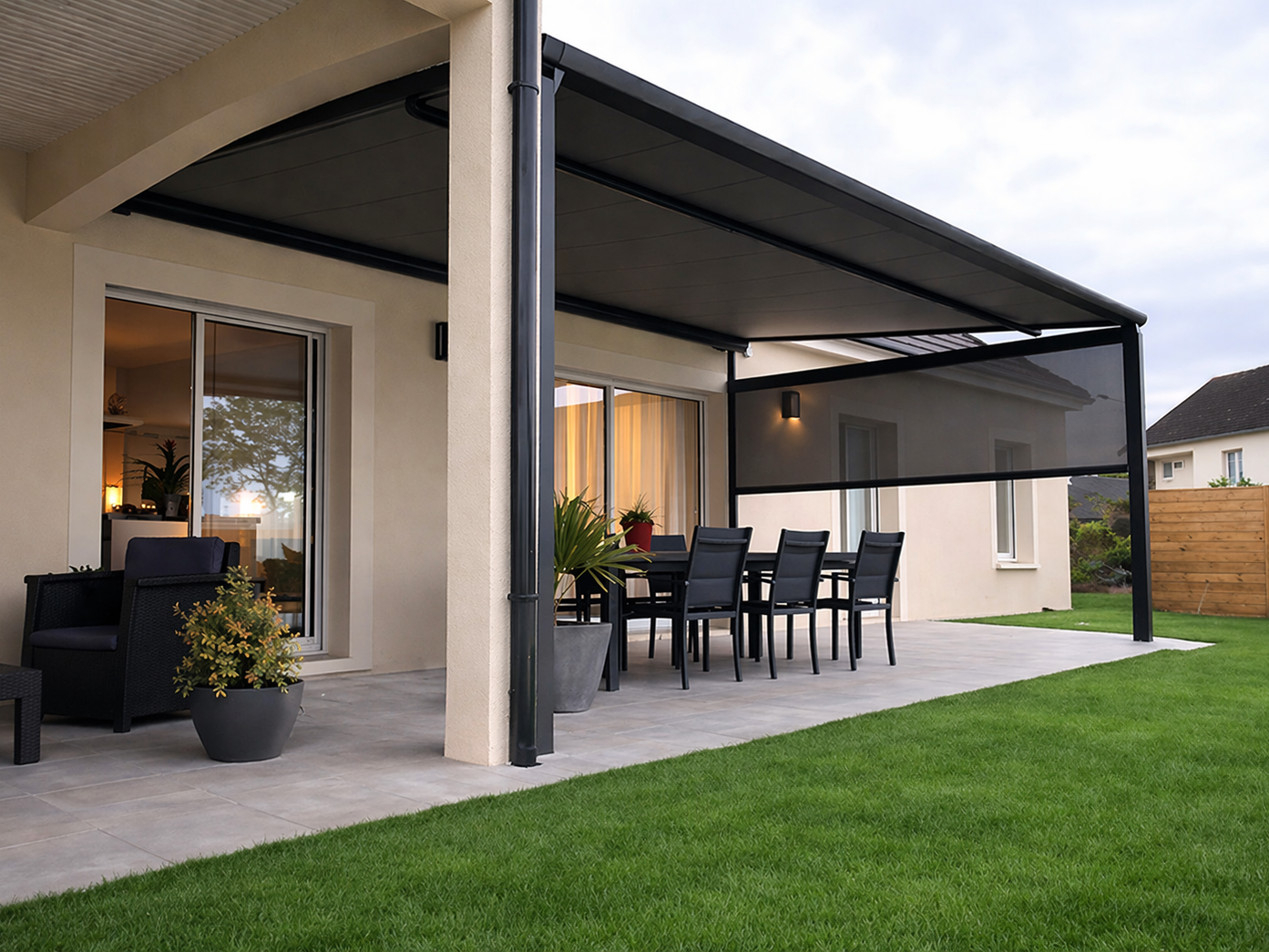 Auvent noir au-dessus d'une terrasse avec table et chaises noires, maison beige et petite clôture en piquets.