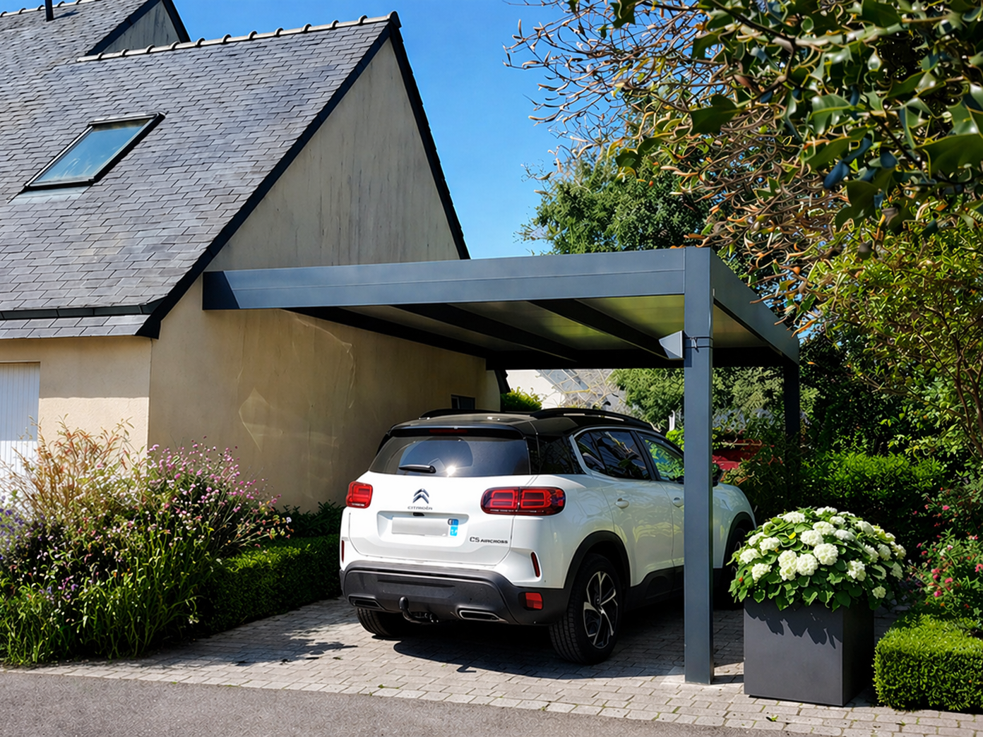 Une voiture argentée est garée sous un abri de voiture gris. Pelouse verte, maison en arrière-plan.