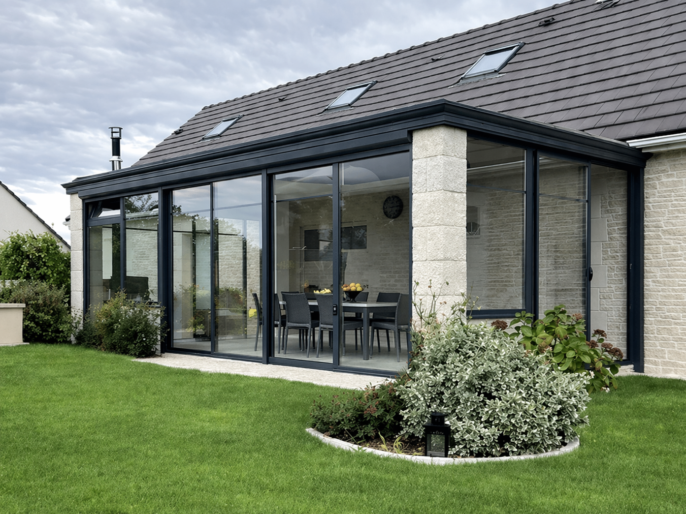 Terrasse arrière avec parois vitrées, verdure et foyer extérieur. Maison au toit gris et aux murs en pierre claire.