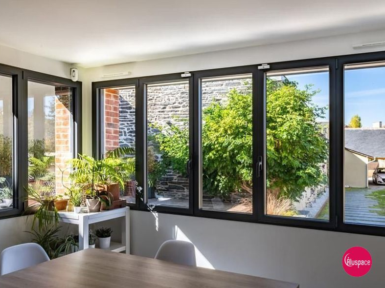 Véranda aux fenêtres à cadre noir, baignée de lumière. Vue sur la verdure extérieure. Table et chaises au premier plan.