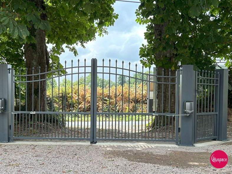 Portail d'allée en métal gris, ouvert, entre deux arbres. Allée en gravier.