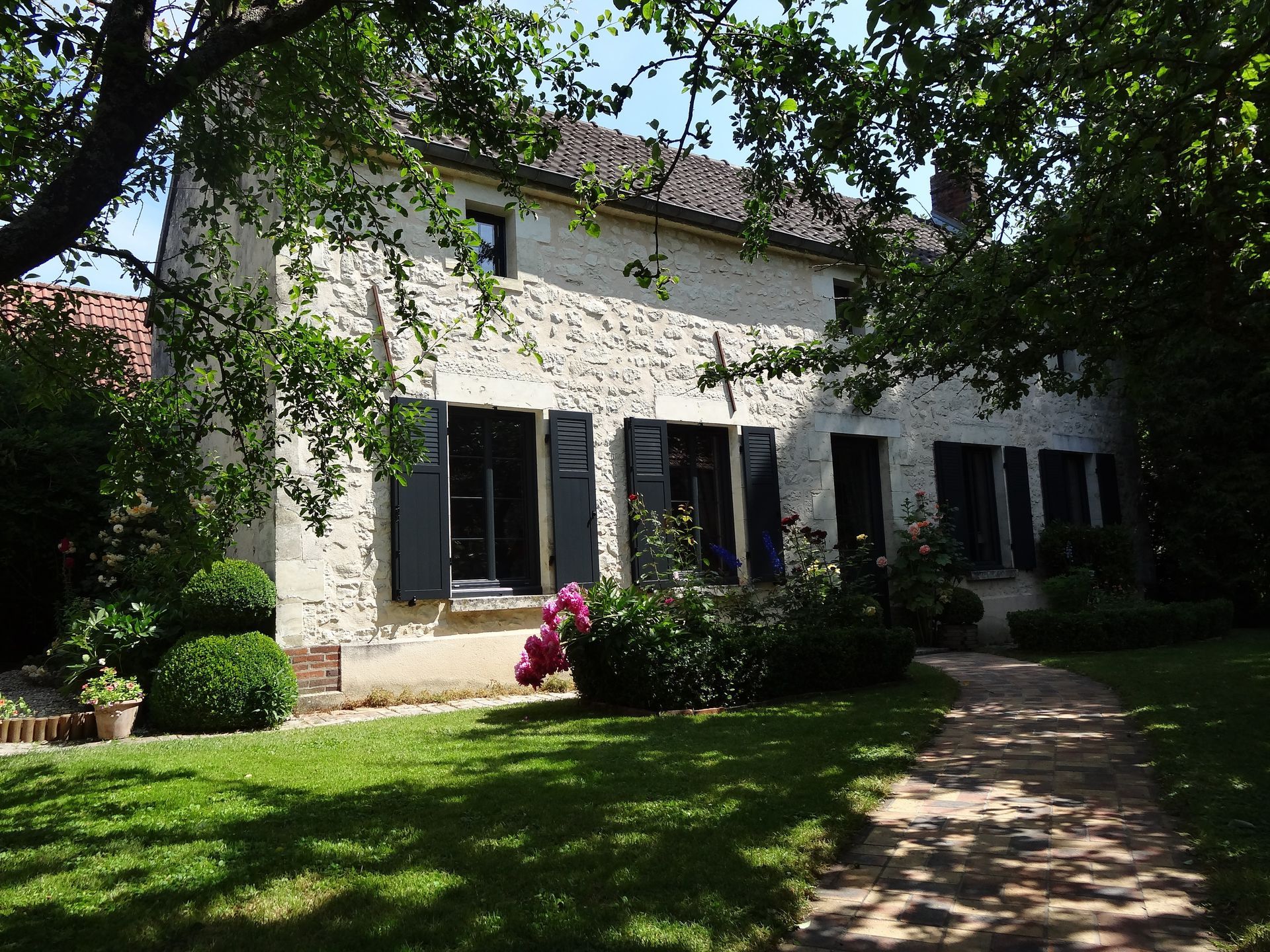 Maison en pierre aux volets sombres, entourée d'herbe verte, d'arbres et de fleurs sous un ciel ensoleillé.