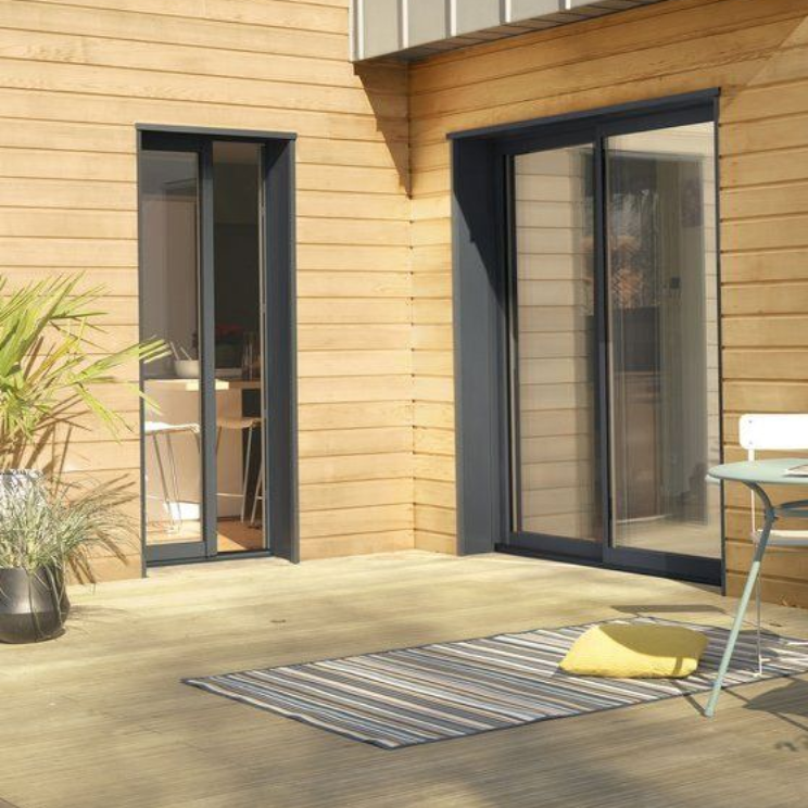 Maison revêtue de bois avec porte-fenêtre coulissante et patio, agrémenté d'un tapis et de mobilier de jardin.