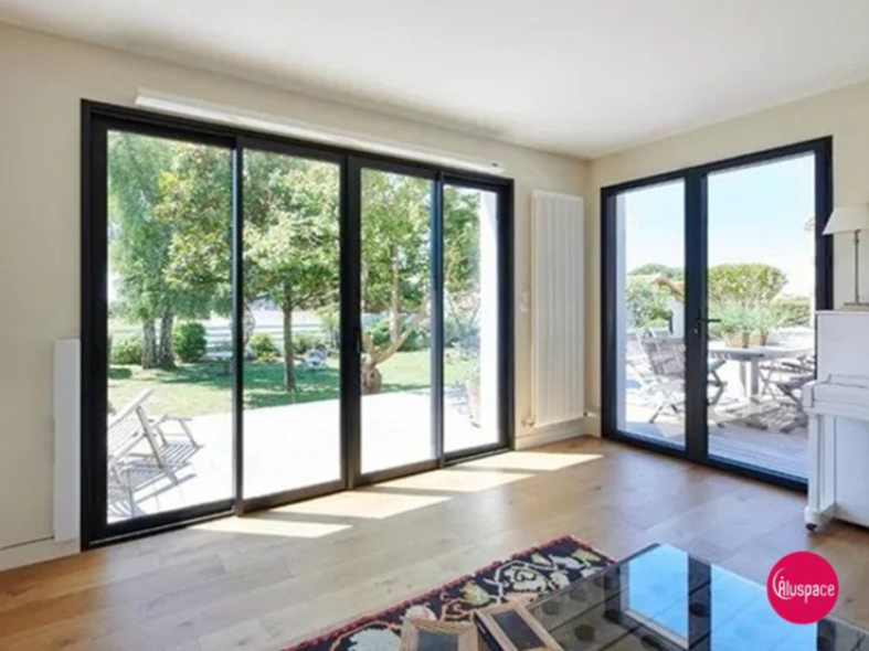 Intérieur lumineux avec portes coulissantes en verre donnant sur une cour. Parquet, encadrements de fenêtres noirs et tapis.