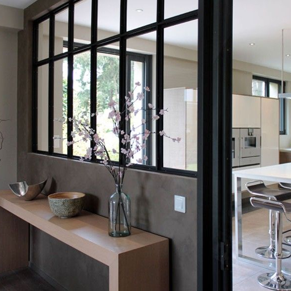 Intérieur avec une baie vitrée à cadre noir, dévoilant une cuisine moderne. Des fleurs dans un vase sont posées sur une étagère.