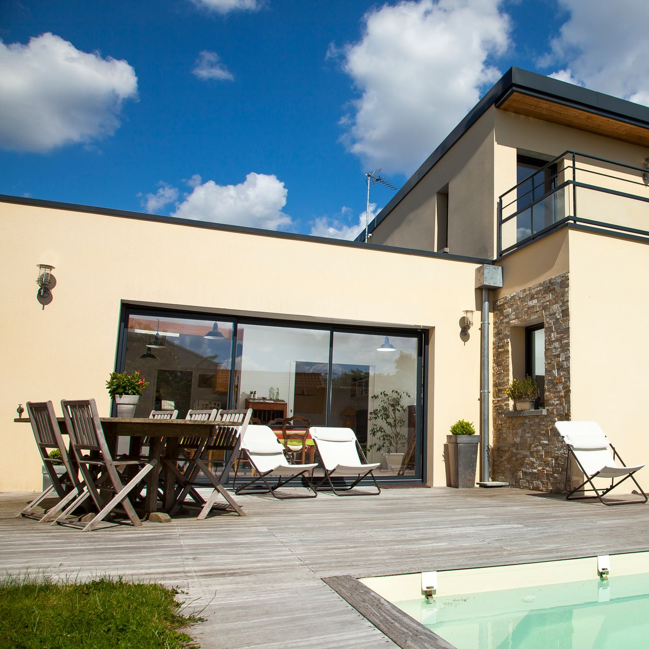 Maison moderne avec terrasse, piscine et grande baie vitrée coulissante. Journée ensoleillée avec ciel bleu et quelques nuages.