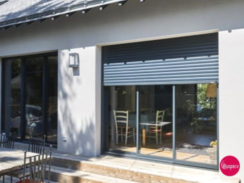 Vue extérieure d'une maison avec de grandes portes vitrées et un volet roulant gris partiellement fermé.
