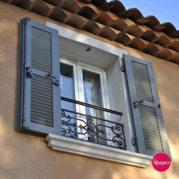 Fenêtre à volets gris, cadre blanc et appui de fenêtre en pierre, contre un mur clair.
