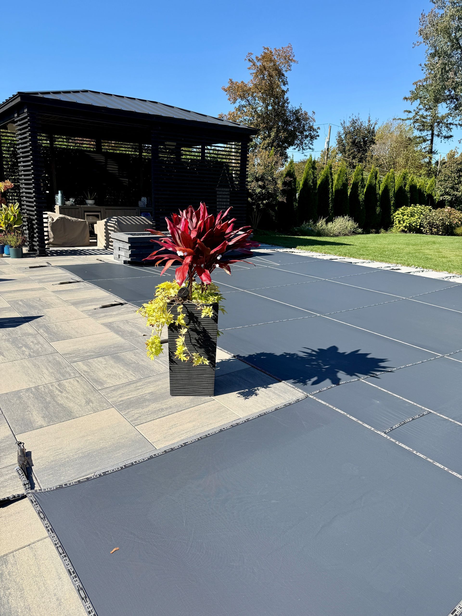 Terrasse avec piscine couverte et pergola ; un pot de fleurs rouge est placé près de la piscine. Journée ensoleillée.
