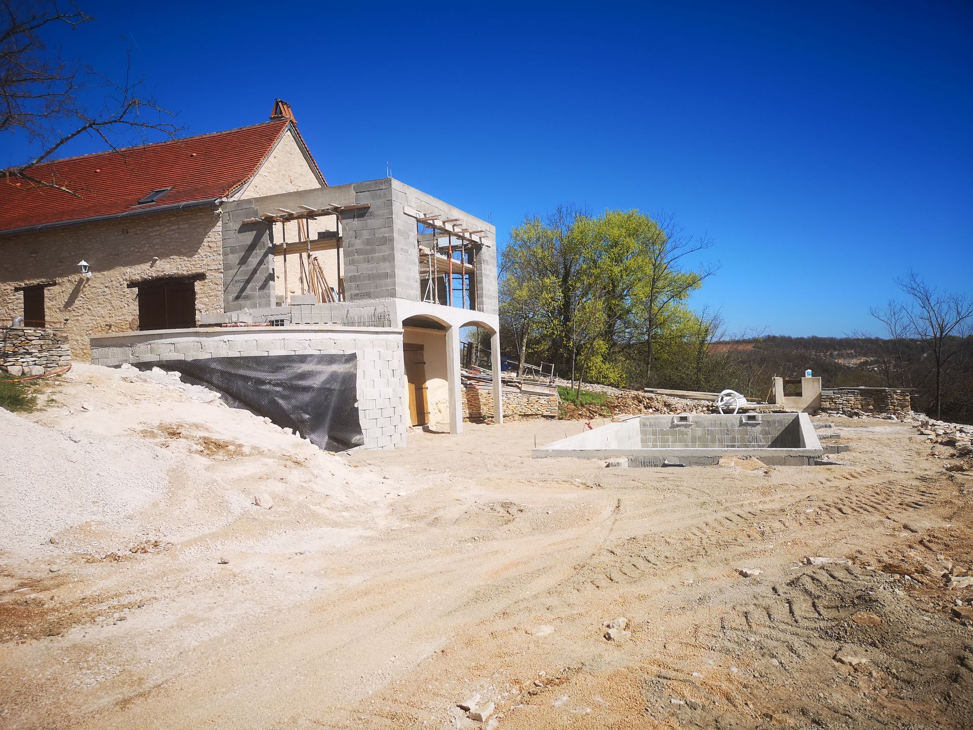 Une maison en construction avec une façade en pierre, une nouvelle extension et une piscine rectangulaire vide.