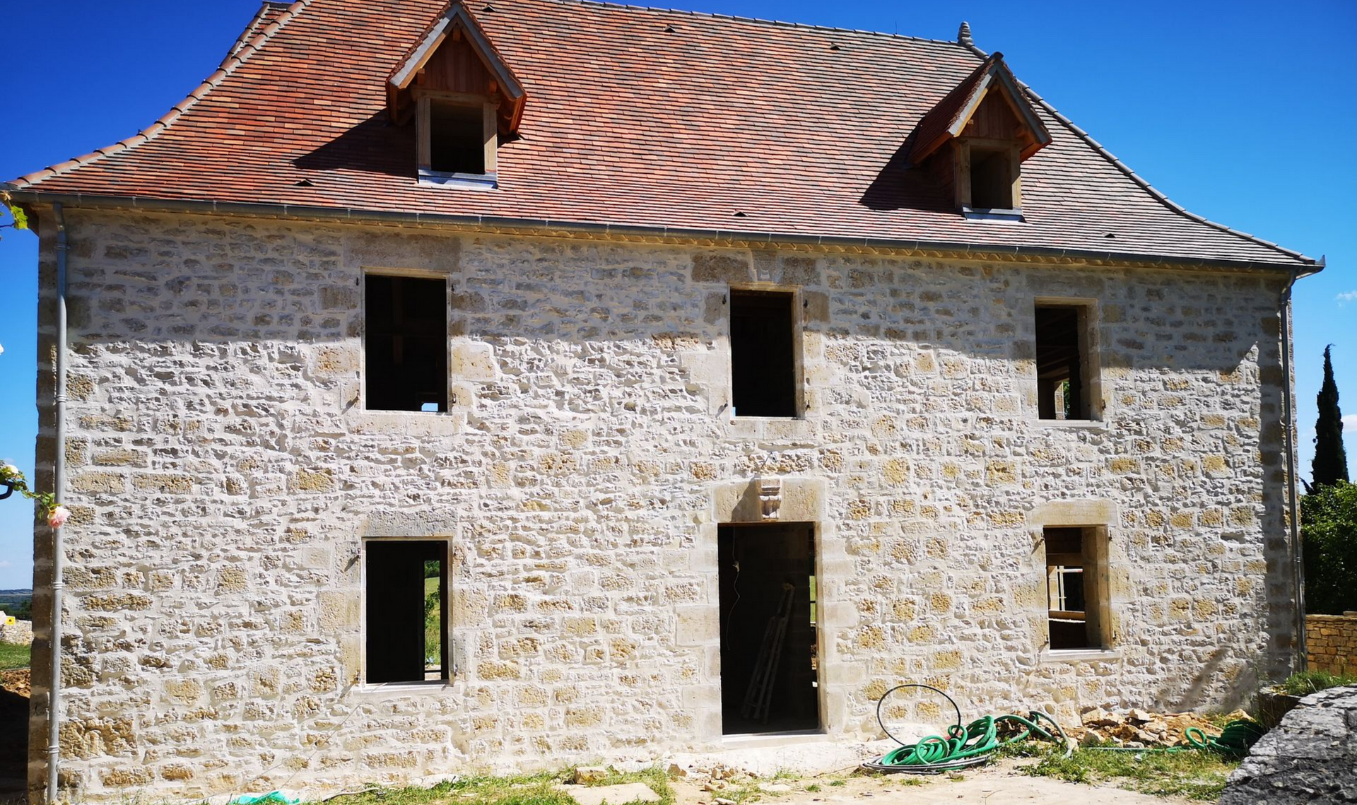 Une maison en pierre à deux étages, avec un toit de tuiles rouges et des cadres de portes et de fenêtres ouverts.