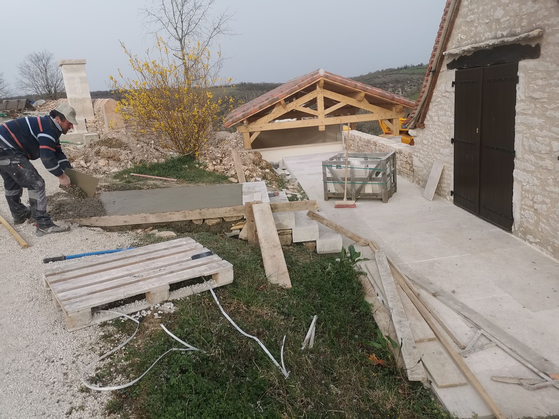 Un ouvrier lisse un chemin en béton fraîchement coulé à côté d'un bâtiment en pierre.