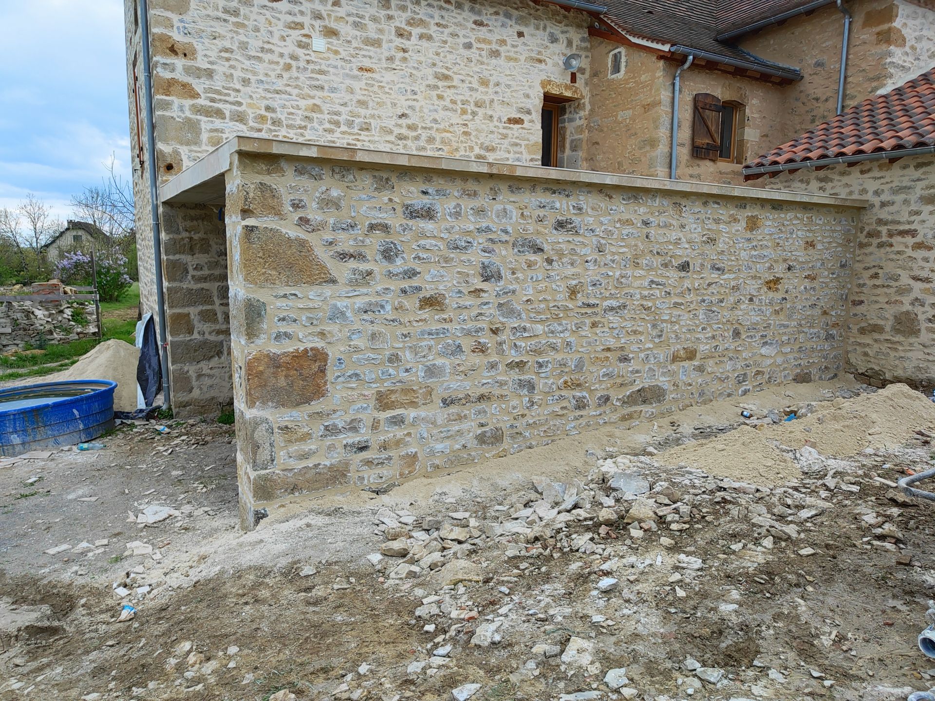 Un mur de pierre nouvellement construit, accolé à un bâtiment rustique en pierre.