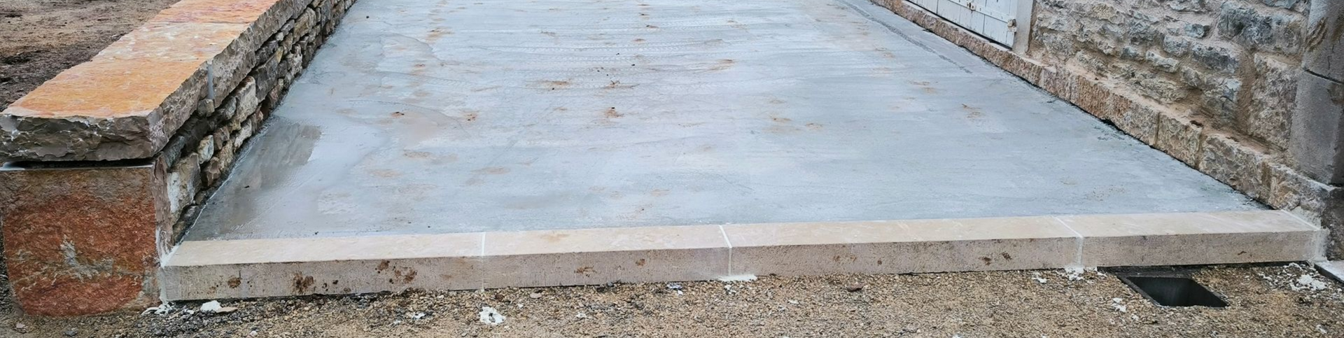Un chemin de pierres plates mène à une terrasse en béton, bordée à gauche par un muret en pierres.