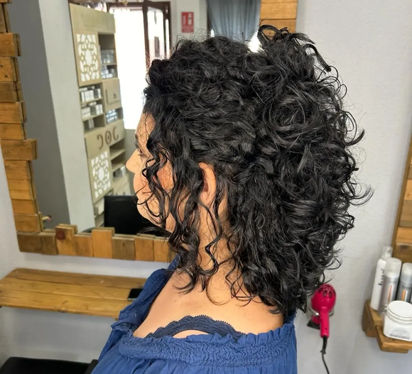 Mujer con cabello oscuro y rizado peinado en un salón, vistiendo una blusa azul.