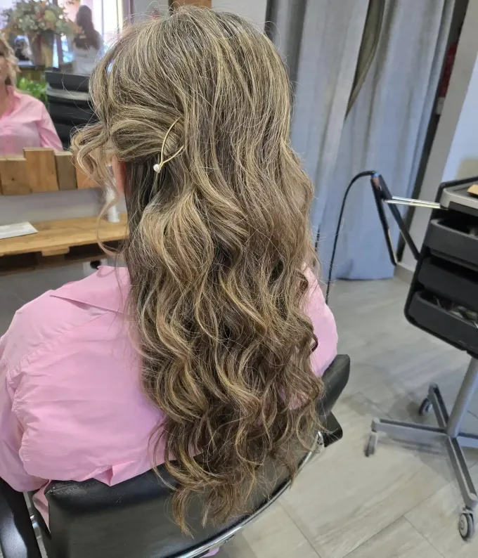 Mujer con cabello largo trenzado peinado en un salón, vistiendo una camisa rosa, sentada en una silla.